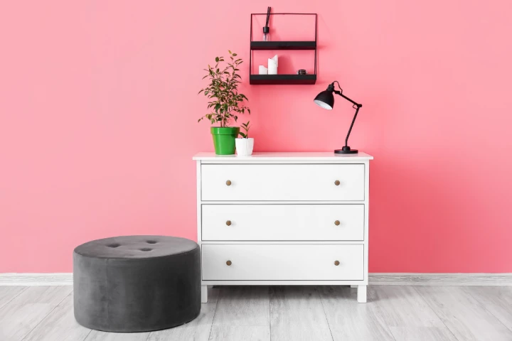 White chest of drawers and black ottoman in front of a pink wall.