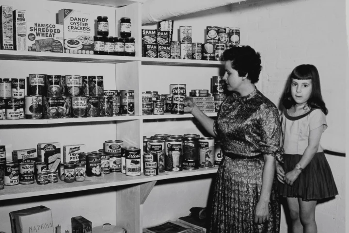A woman and a young girl in a pantry, looking at canned goods and boxes on shelves.