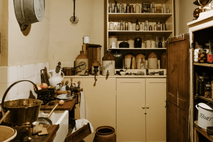 Vintage kitchen with wooden cupboards, cluttered countertop filled with utensils and jars, and shelves stocked with old containers and crockery.